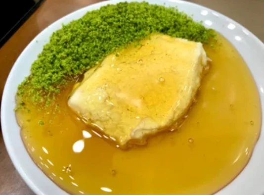 A piece of kaymak sitting in honey on a plate, with ground pistachio alongside