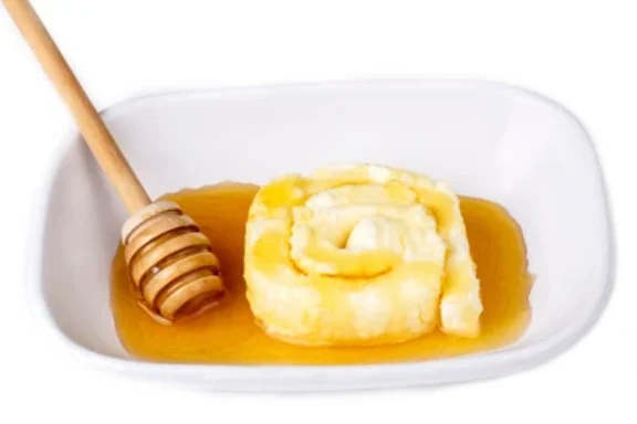 A roll of kaymak on a white dish with honey poured around it, and a wooden honey dipper beside